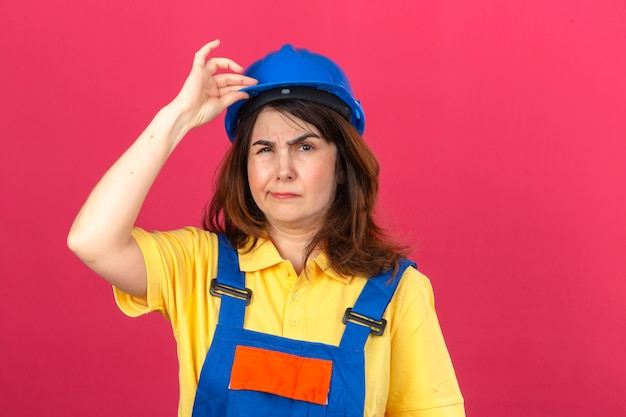 Constructor mujer vestida con uniforme de construcción y casco de seguridad con expresión escéptica tocando casco parado sobre pared rosa aislado