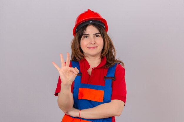 Constructor mujer vestida con uniforme de construcción y casco de seguridad sonriendo amigable mostrando y apuntando hacia arriba con los dedos número tres sobre la pared blanca aislada