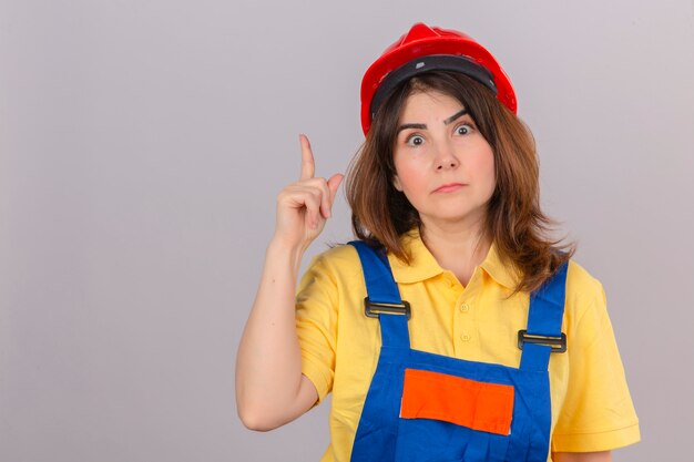 Constructor mujer vistiendo uniforme de construcción y casco de seguridad mirando sorprendido apuntando hacia arriba con el dedo de pie sobre la pared blanca aislada