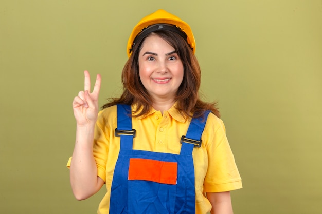 Constructor mujer vistiendo uniforme de construcción y casco de seguridad mostrando y apuntando hacia arriba con los dedos número dos sonriendo alegre de pie sobre la pared verde aislado