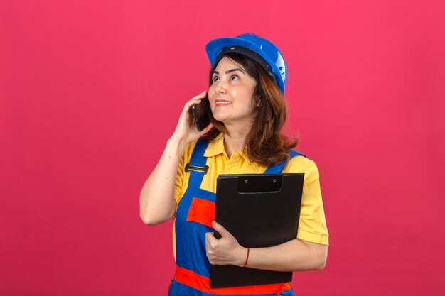 Constructor mujer vistiendo uniforme de construcción y casco de seguridad con portapapeles mientras habla por teléfono móvil mirando hacia arriba con una sonrisa en la cara sobre la pared rosa aislado