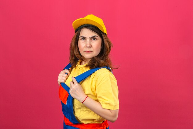 Foto gratuita constructor mujer vistiendo uniforme de construcción y gorra amarilla con el ceño fruncido disgustado de pie sobre la pared rosa aislado