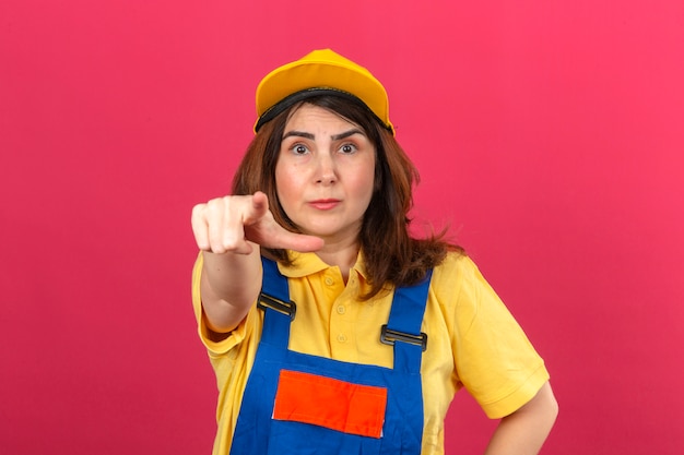 Constructor mujer vistiendo uniforme de construcción y gorra amarilla mirando sorprendido señalando con el dedo a la cámara sobre la pared rosa aislada