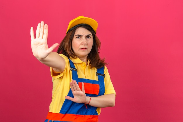 Constructor mujer vistiendo uniforme de construcción y gorra amarilla de pie con las manos abiertas haciendo señal de stop con gesto de defensa de expresión seria y segura sobre pared rosa aislado