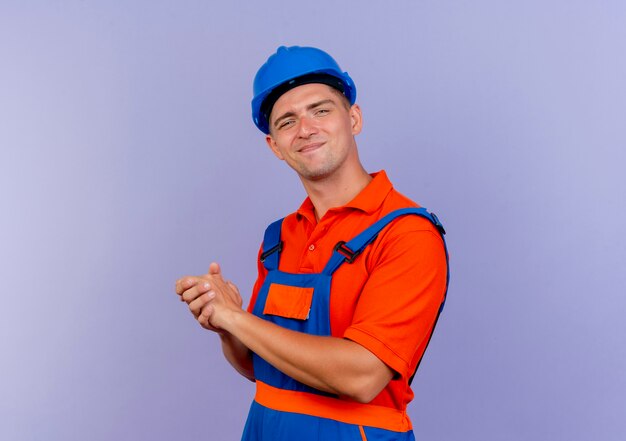 Constructor de sexo masculino joven complacido vistiendo uniforme y casco de seguridad mostrando gesto de apretones de manos en púrpura