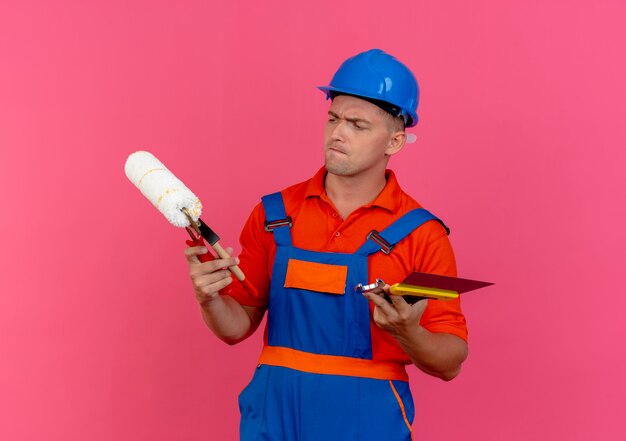 Constructor de sexo masculino joven confundido vistiendo uniforme y casco de seguridad sosteniendo y mirando herramientas de construcción