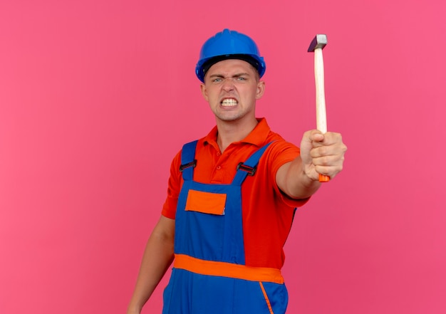 Constructor de sexo masculino joven enojado vistiendo uniforme y casco de seguridad sosteniendo el martillo