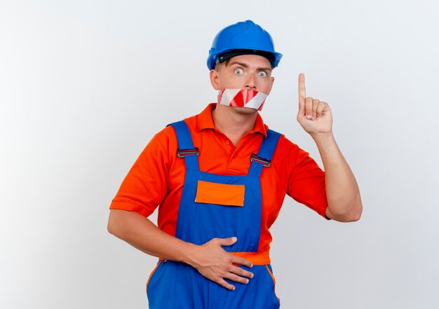 Constructor de sexo masculino joven sorprendido con uniforme y casco de seguridad selló su boca con cinta adhesiva y apunta hacia arriba