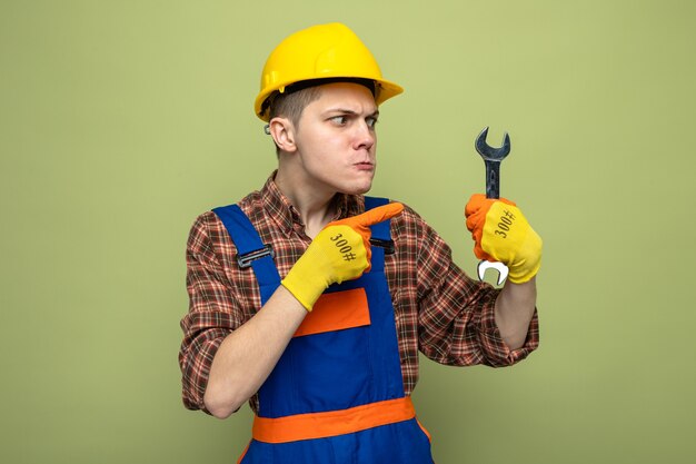 Foto gratuita constructor de sexo masculino joven vistiendo uniforme con guantes sosteniendo y puntos en la llave de boca aislada en la pared verde oliva