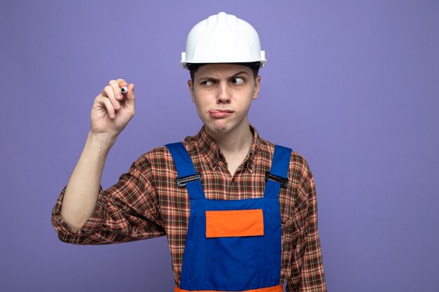 Constructor de sexo masculino joven vistiendo uniforme sosteniendo y mirando el marcador aislado en la pared púrpura
