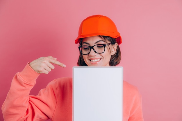 Constructor sonriente del trabajador de la mujer sostenga el tablero blanco de la muestra en blanco sobre fondo rosa. Casco de construcción. Señalar con el dedo índice.