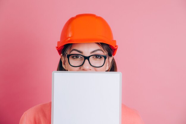 Constructor sonriente del trabajador de la mujer sostenga el tablero blanco de la muestra en blanco sobre fondo rosa. Casco de construcción.