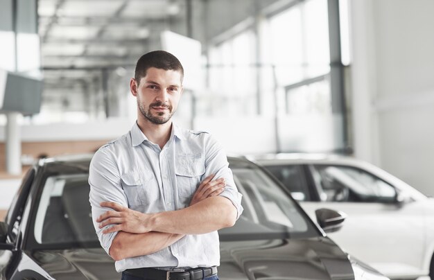 Consultor de joven guapo en el salón del coche de pie cerca del coche.