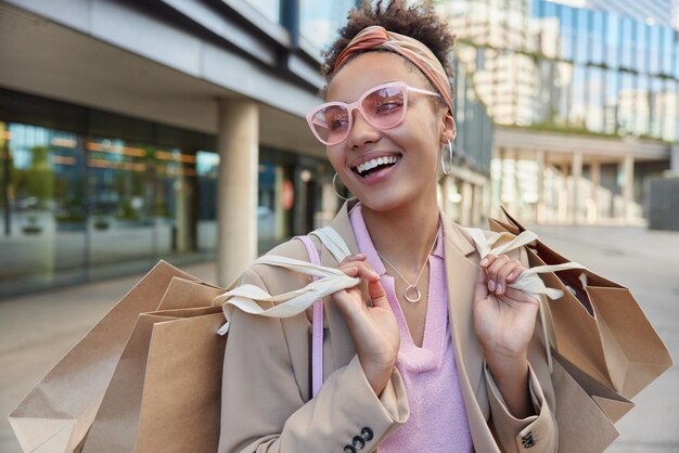 El consumismo de la gente y el concepto de compra Una mujer de moda alegre usa gafas de sol rosas, una elegante chaqueta lleva bolsas de papel y tiene un estado de ánimo feliz, paseos en la calle contra el fondo urbano