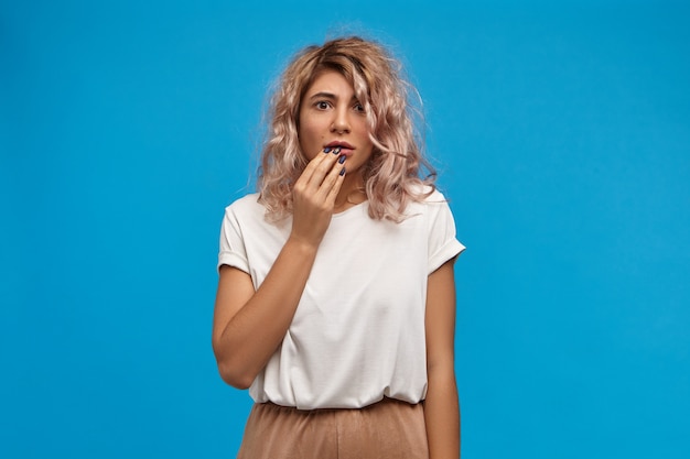 Contador de mujer joven preocupada emocional con peinado elegante posando contra el espacio de la pared azul en blanco, tocando los labios, siendo nervioso