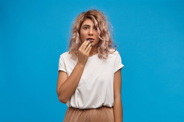 Contador de mujer joven preocupada emocional con peinado elegante posando contra el espacio de la pared azul en blanco, tocando los labios, siendo nervioso