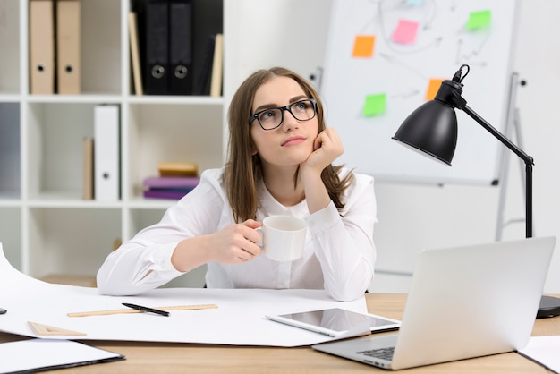 Contemplado arquitecto mujer sosteniendo una taza de café sentado en el lugar de trabajo