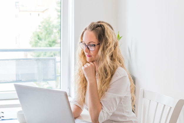 Contemplado rubia mujer joven sentada cerca de la ventana con la computadora portátil