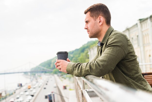 Contemplando al hombre joven que sostiene la taza de café disponible que mira la vista de la ciudad