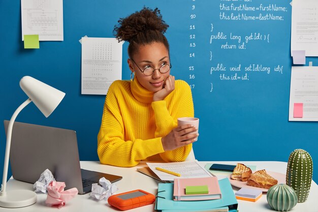 Contemplativa mujer afroamericana independiente bebe cafeína de la taza, mira pensativamente a un lado, vestida con un suéter amarillo, usa laptop