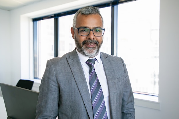 Contenido CEO indio de pie y sonriendo para retrato. Exitoso empresario barbudo pensativo en gafas posando en la sala de la oficina. Concepto de negocio, expresión y gestión