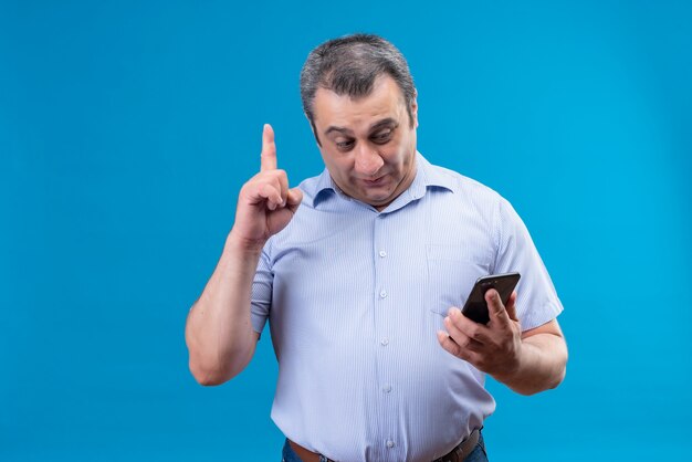 Contenido hombre de mediana edad en camisa de rayas azules mirando el teléfono móvil para encontrar una excelente solución a su problema en un espacio azul