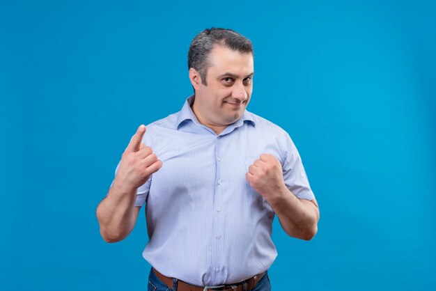 Contenido hombre de mediana edad en camisa a rayas sosteniendo el puño cerrado y apuntando con el dedo índice en un espacio azul