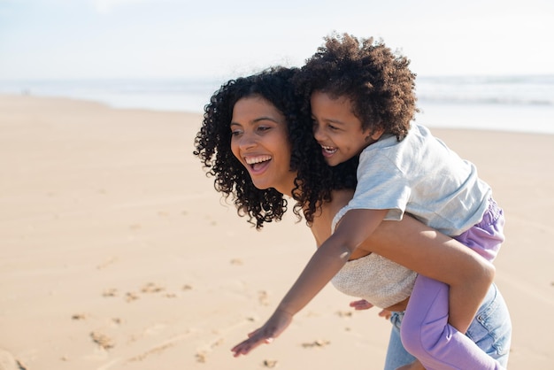 Contenido madre e hija pasando tiempo en la playa. Familia afroamericana caminando, riendo, jugando, montando en la espalda. Ocio, tiempo en familia, concepto de paternidad.