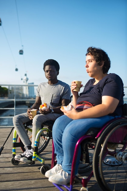 Contenido pareja birracial bebiendo café en un día soleado. Hombre afroamericano y mujer caucásica en sillas de ruedas en el terraplén, bebiendo bebidas calientes de tazas. Merienda, relación, concepto de felicidad.