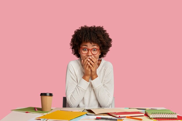 El contenido sorprendió a una mujer negra que se tapa la boca, mira con alegría, no puede creer en la promoción, tiene una reacción complacida sobre algo positivo, se sienta en el escritorio con papelería. Estudiantes llenos de alegría estudian en el interior