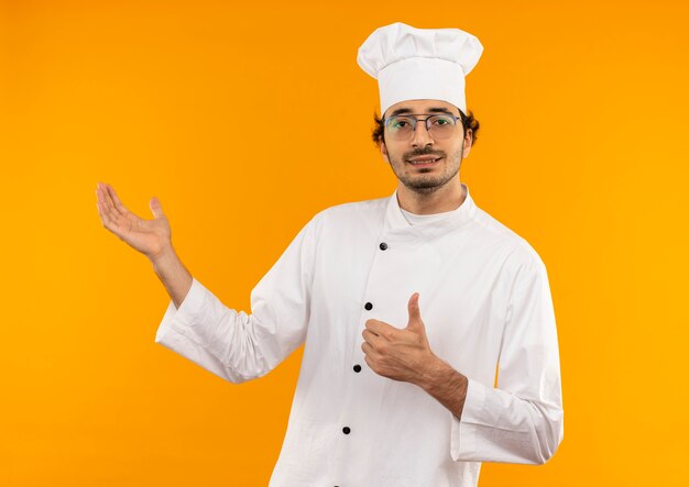 Contento joven cocinero vistiendo uniforme de chef y puntos de gafas con la mano al lado de su pulgar hacia arriba