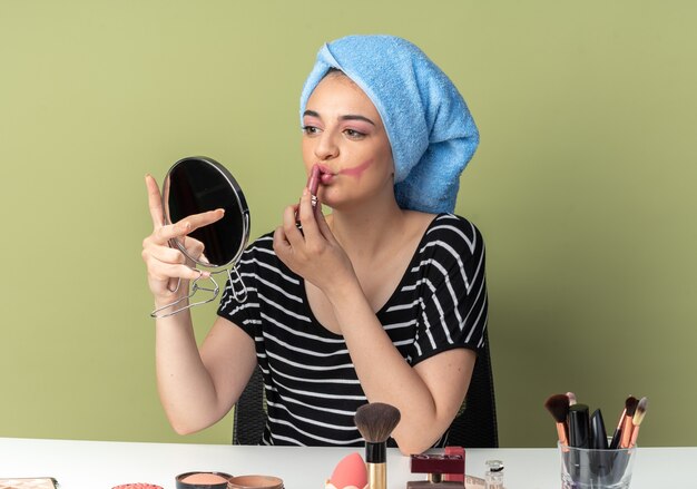 Contento mirando al espejo hermosa joven se sienta a la mesa con herramientas de maquillaje envuelto el pelo en una toalla aplicando lápiz labial aislado en la pared verde oliva