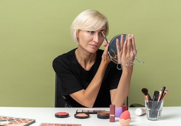 Contento mirando al espejo joven hermosa se sienta a la mesa con herramientas de maquillaje aplicando sombra de ojos aislada en la pared verde oliva