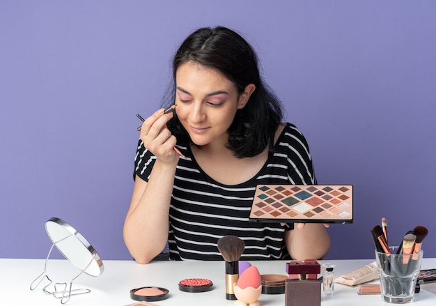 Contento mirando al espejo joven hermosa se sienta a la mesa con herramientas de maquillaje aplicando sombra de ojos con pincel de maquillaje aislado en la pared azul