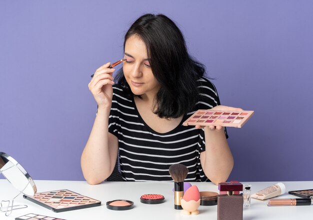 Contento mirando al espejo joven hermosa se sienta a la mesa con herramientas de maquillaje aplicando sombra de ojos con pincel de maquillaje aislado en la pared azul