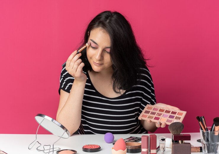 Foto gratuito contento mirando al espejo joven hermosa se sienta a la mesa con herramientas de maquillaje aplicando sombra de ojos con pincel de maquillaje aislado en la pared rosa