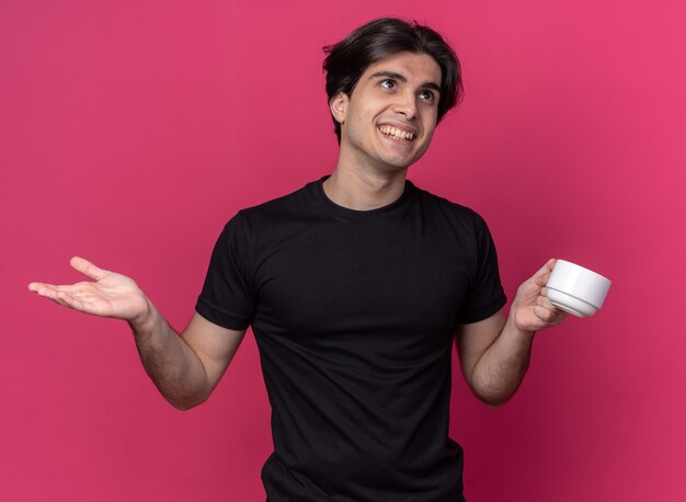 Contento mirando al lado chico guapo joven con camiseta negra sosteniendo una taza de café extendiendo la mano aislada en la pared rosa