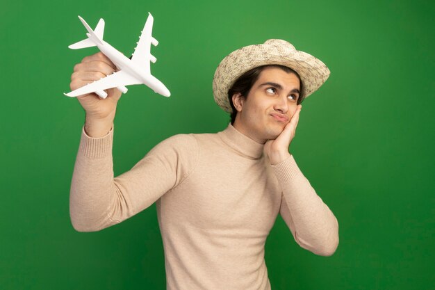 Foto gratuita contento mirando al lado chico guapo joven con sombrero y levantando avión de juguete poniendo la mano en la mejilla aislada en la pared verde