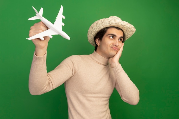 Contento mirando al lado chico guapo joven con sombrero y levantando avión de juguete poniendo la mano en la mejilla aislada en la pared verde