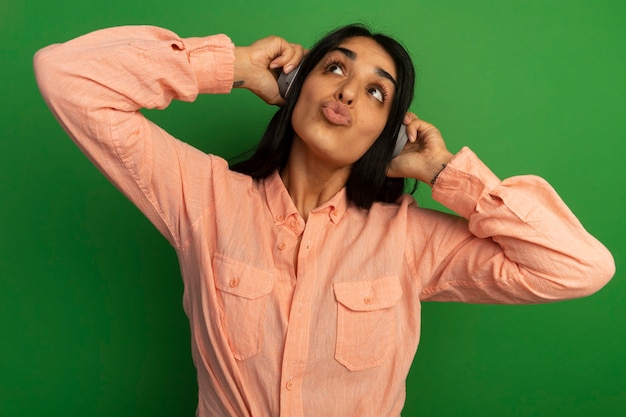 Contento mirando al lado hermosa joven vestida con camiseta rosa con auriculares aislados en la pared verde