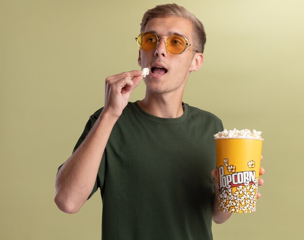 Contento mirando al lado joven guapo con camisa verde con gafas sosteniendo y probando un cubo de palomitas de maíz aislado en la pared verde oliva