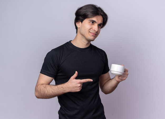 Contento mirando al lado joven guapo con camiseta negra sosteniendo y apunta a una taza de café aislada en la pared blanca