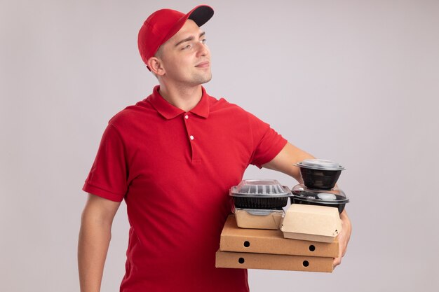 Contento mirando al lado joven repartidor con uniforme con gorra sosteniendo contenedores de comida en cajas de pizza aisladas en la pared blanca