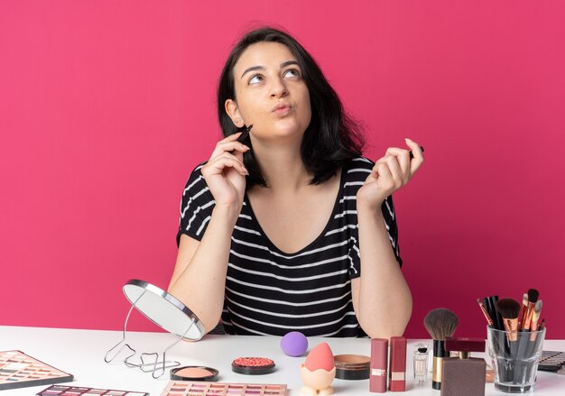 Contento mirando hacia arriba la hermosa joven se sienta a la mesa con herramientas de maquillaje con delineador de ojos aislado en la pared rosa