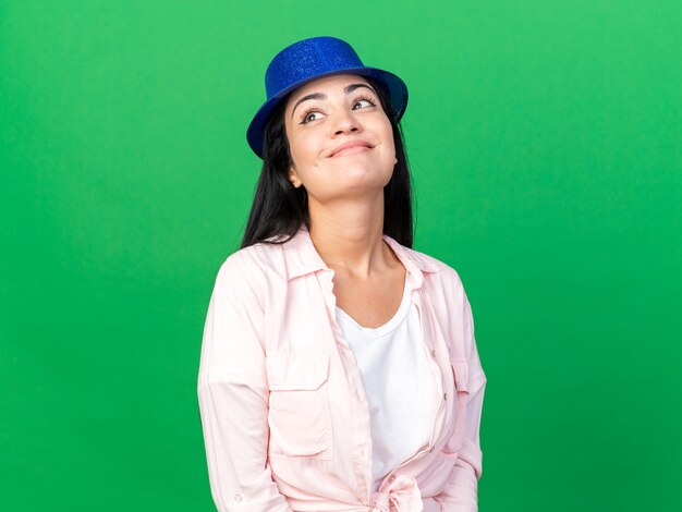 Contento mirando hacia arriba joven hermosa mujer vistiendo gorro de fiesta aislado en la pared verde