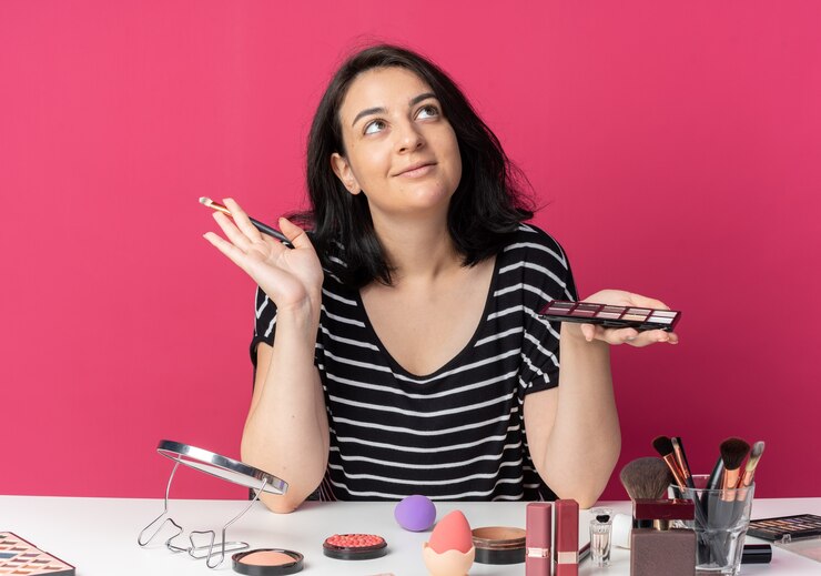 Foto gratuito contento mirando hacia arriba joven hermosa se sienta a la mesa con herramientas de maquillaje sosteniendo la paleta de sombra de ojos con pincel de maquillaje aislado en la pared rosa