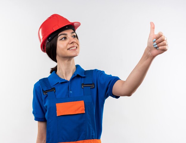 Contento mirando a la mujer joven constructora de lado en uniforme mostrando el pulgar hacia arriba