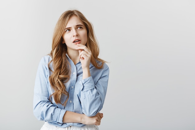 Continúe con la dieta o pida pizza. Retrato de mujer atractiva de pensamiento cuestionado con cabello rubio y rizos, tocando el labio y mirando hacia arriba con expresión reflexiva, teniendo idea sobre la pared gris