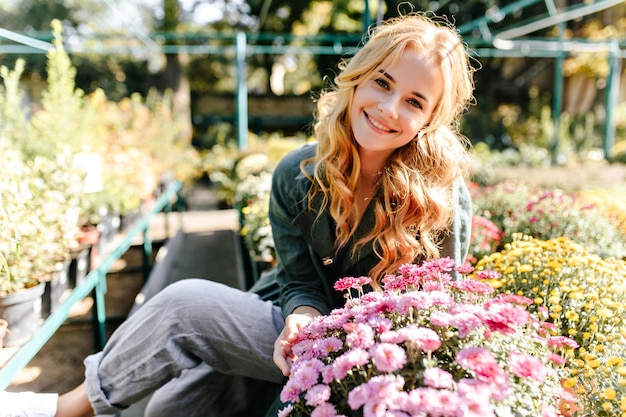 Contra el fondo de plantas con flores brillantes y arbustos verdes que crecen en el jardín, una hermosa chica rubia con ojos verdes sonríe dulcemente y disfruta del sol