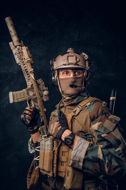 Contratista de servicios de seguridad privada con uniforme de camuflaje posando con rifle de asalto. Foto de estudio contra una pared de textura oscura